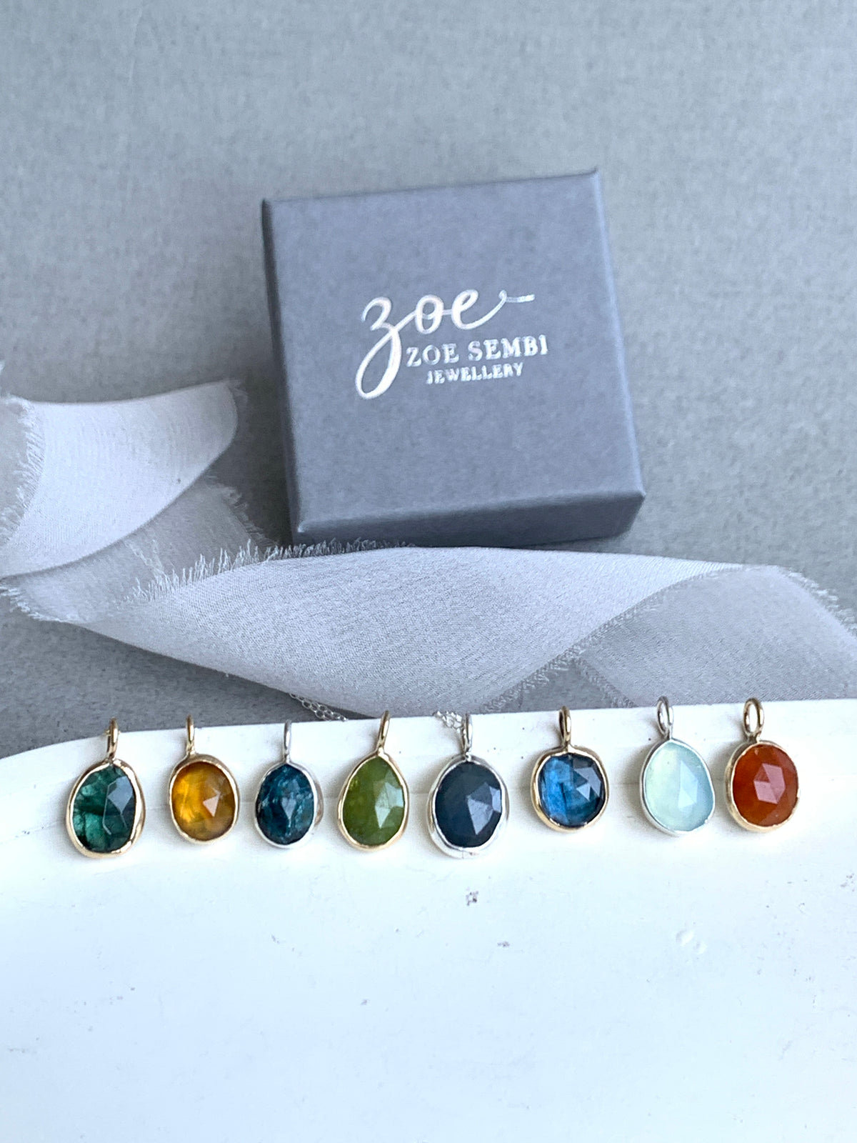 A set of seven colorful gemstone drop earrings in gold settings, displayed next to a gray jewelry box with the brand name 'Zoe Sembi'. The earrings feature round cabochon stones in orange, light blue, dark blue, olive green, dark teal, yellow, and emerald green, suspended from gold rings.