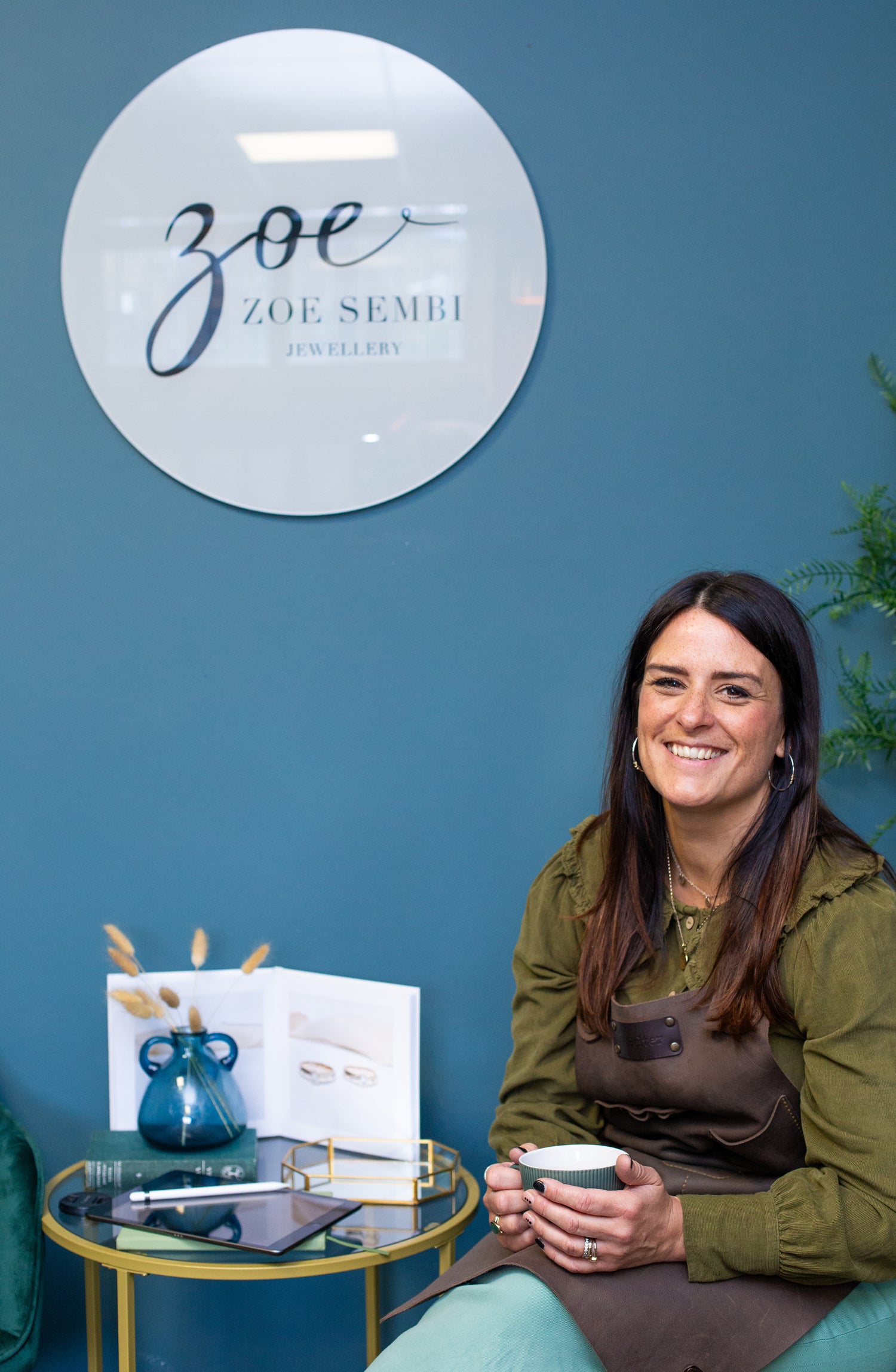 Woman sitting in front of a 'Zoe Semli Jewellery' sign, holding a cup.