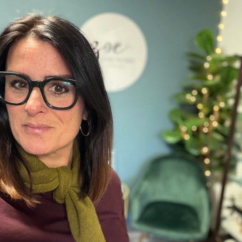 Woman wearing glasses and a green scarf in a room with a plant and chair.